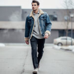 Homme marchant dans une rue en ville en hiver, vêtu d’une veste en jean bleu brut fourrée beige, sous une lumière naturelle.