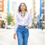 Femme élégante en jean et chemise blanche, portant des accessoires colorés dans une rue urbaine lumineuse.