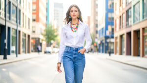 Femme élégante en jean et chemise blanche, portant des accessoires colorés dans une rue urbaine lumineuse.