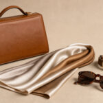 Sélection élégante d’accessoires féminins intemporels composée d’un sac, d’un foulard en soie, d’une montre classique et de lunettes sur un fond beige neutre.